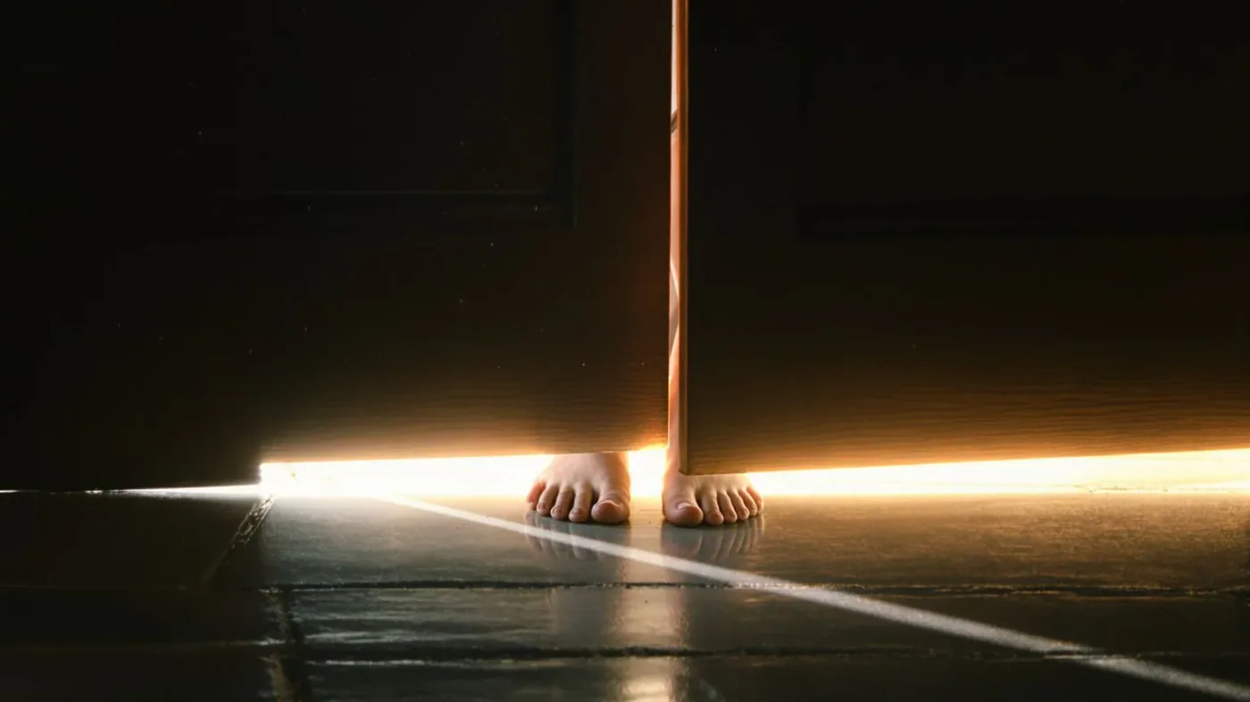 Bare feet at a doorway threshold where warm light meets shadow showing what changes and softens first