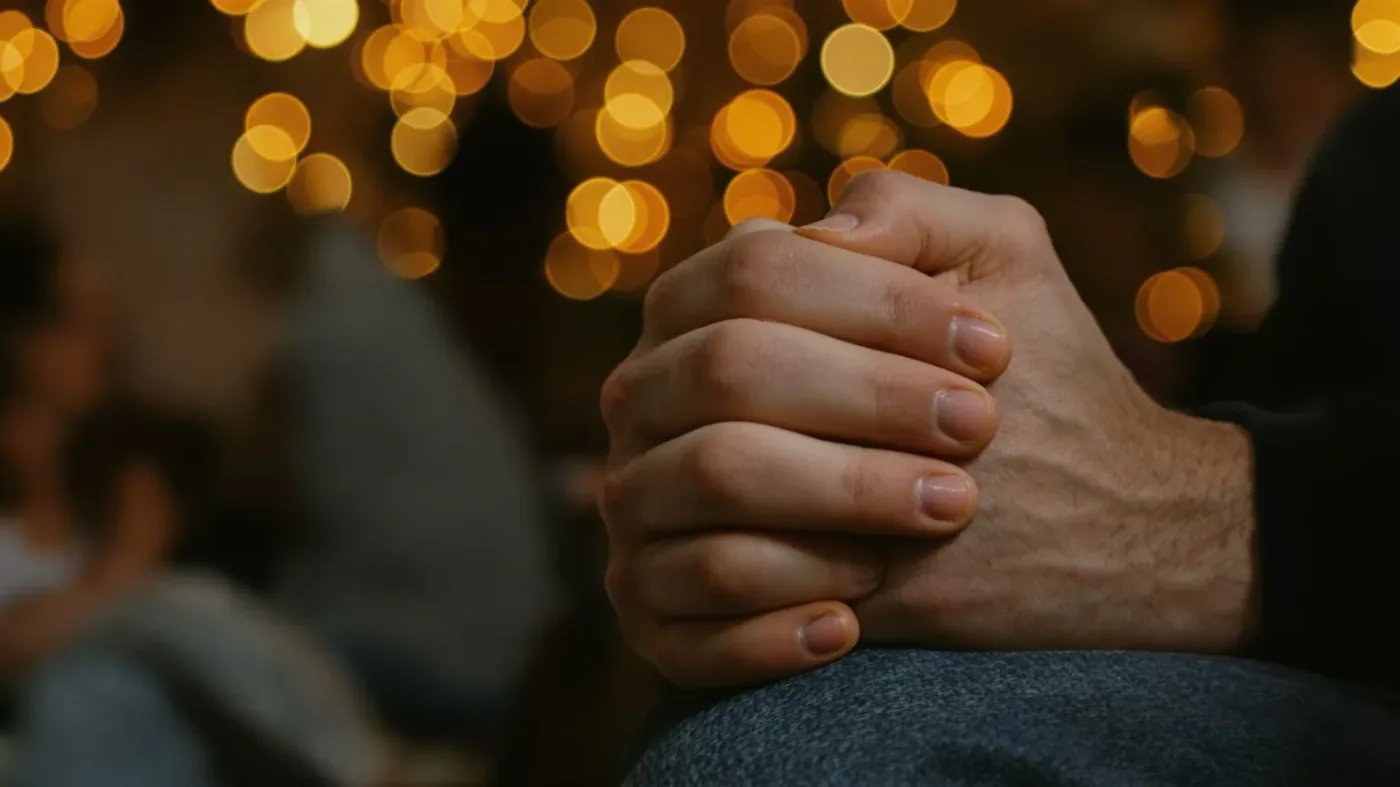 Close-up of tense clasped hands in lap while people gather in background feeling empty
