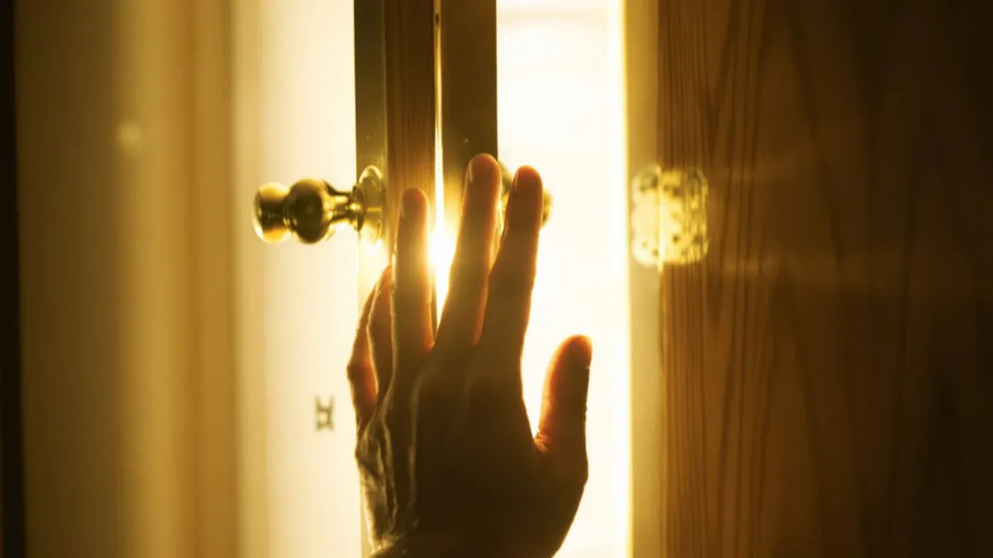 Hesitant hand reaching toward a partially open door with warm light streaming through the gap showing fear of being real