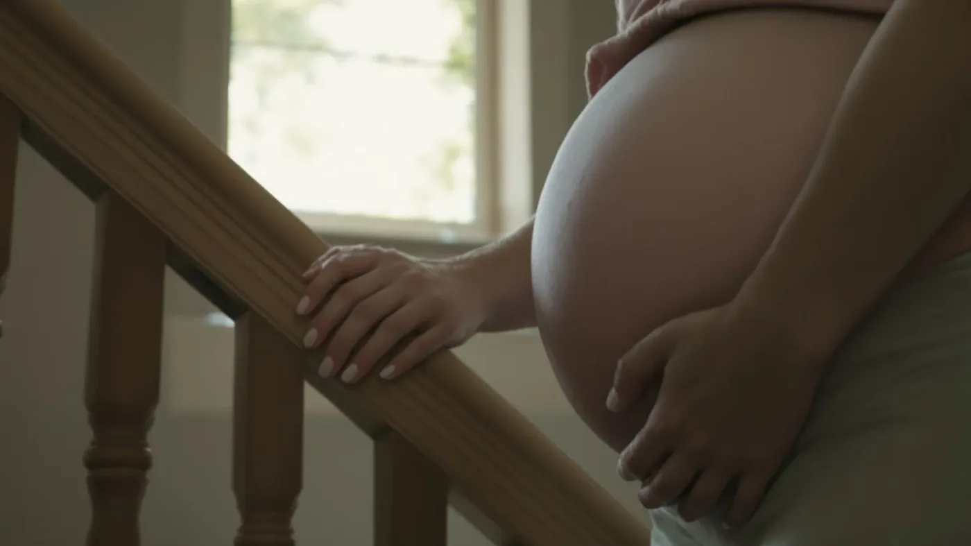 Pregnant woman's tense hands gripping a wooden staircase railing, body paused mid-step