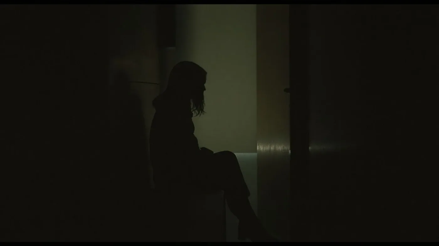 Woman gripping bench edge in dim hallway facing lit doorway showing why emotional masking feels safer than honesty