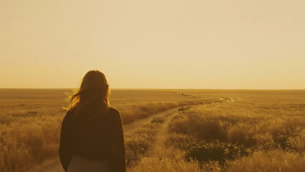 Woman standing at edge of vast golden grassland looking toward horizon path, evoking why cant i cry emotional stillness